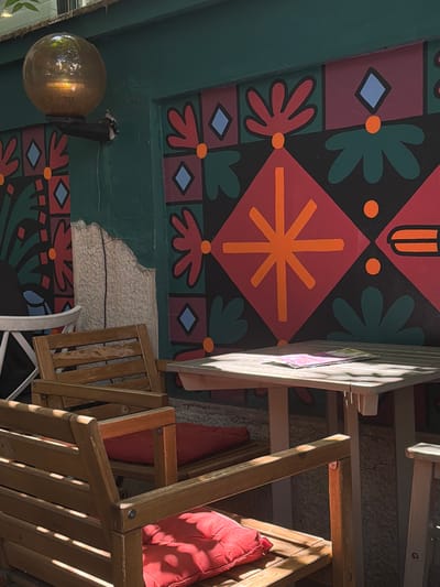 The image depicts an outdoor seating area with a colorful mural as a backdrop. In the foreground, two wooden chairs with red cushions are visible. To the right, a wooden table holds a couple of papers, and a portion of another chair can be seen. Behind the seating area, a green wall features a vibrant mural with geometric patterns and floral motifs in red, black, green, and blue. A spherical lamp with a warm light source is mounted on the wall above the mural. The environment appears to be an urban setting, possibly a cafe or restaurant patio. The image does not show any people. There is no information available about the specific location in Vracar Urban Municipality, Serbia.