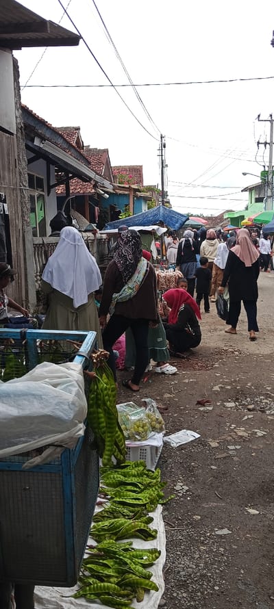 The scene takes place on a street in Kutawaringin, Indonesia, with the coordinates -6.974 latitude and 107.519 longitude. Several people are visible, some dressed in traditional clothing, such as headscarves. There are several objects in the scene, including a blue metal cage filled with green vegetable pods arranged on a white cloth, a bag filled with the same pods, a bag of small round fruit, and a few other bags. Several people are walking around, likely buying and selling these items, while others stand, wait, or sit. Several buildings line the street and there are overhead power lines. The street is paved with a mix of gray gravel and pavement.
