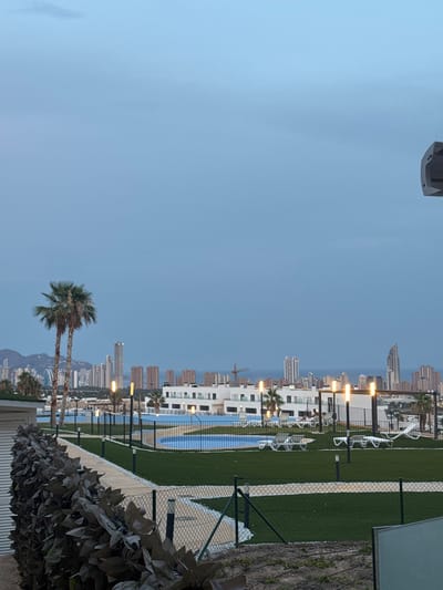 The image depicts a twilight view of a modern residential complex with a swimming pool and meticulously manicured green spaces. In the foreground, a textured dark green hedge runs along a fence. Beyond the hedge, a paved walkway curves around a bright blue swimming pool and an expanse of artificial turf dotted with lounge chairs. Several modern lamp posts with illuminated bulbs line the walkway. In the mid-ground, a white, multi-story building with balconies is visible. In the background, a dense cityscape with numerous high-rise buildings stretches across the horizon, with mountains visible to the left. The sea is present in the distance. The sky is a pale, overcast blue, suggesting either dawn or dusk. The overall scene is calm and serene. This is likely a resort or residential area located in or near Finestrat, Spain, given the architecture and coastal setting often found in that region.