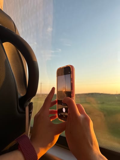 A person is holding a smartphone, photographing a landscape viewed through a window. The landscape includes a sky with a gradient of colors transitioning from blue to orange, likely at sunrise or sunset, and a field. The smartphone is an iPhone with a pink case, displaying the same landscape on its screen. The left side of a car steering wheel is in the foreground. The person's hands and forearm are visible, wearing a burgundy smartwatch band. The location is Bol'shiye Klyuchishchi, Russia, as evidenced by the landscape and metadata from similar images.