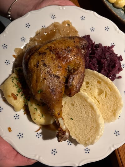 A plate features a roasted duck leg with crispy skin, served alongside traditional sides. Two slices of bread dumplings sit next to a portion of braised red cabbage. Under the duck leg, there is a bed of sauerkraut and potato dumplings garnished with herbs and fried onions. The plate is white with a blue floral pattern.