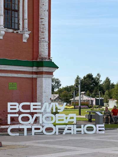 The image depicts an outdoor scene in what appears to be a public space. Dominating the foreground is a large, three-line sign constructed from white, three-dimensional letters. The text on the sign is in Cyrillic script and reads "ВСЕМУ ГОЛОВА СТРОГА" (translation: "Head of everything strogan"). To the left of the sign, a portion of a red brick building with white architectural accents and a green roof is visible. A sign in Russian, indicating cultural heritage, is affixed to the brick wall. In the background, there is a park-like setting with trees, lampposts, benches, and small structures, possibly kiosks or pavilions. Two individuals are seated on a bench in the middle ground, observing the scene. The sky is clear and blue. This location is identified as the Stroganov House in Berezniki, Russia.