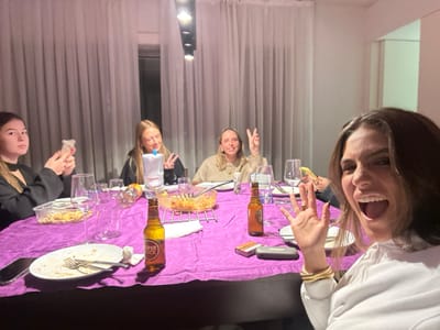 The image depicts an indoor social gathering of four young adult females around a purple-draped dining table, situated in Lordelo do Ouro e Massarelos, Portugal. The woman in the immediate foreground, positioned to the right, has dark hair, wears a white top with gold bracelets, and exhibits an open-mouthed, animated expression while raising her right hand with palm outward towards the camera. Seated left of center at the table, a blonde-haired woman in a dark top forms a peace sign with her right hand. Behind her, another blonde-haired woman, wearing a light-colored top, also makes a peace sign. On the far left of the table, a dark-haired woman in a black top looks left, holding a small white object in her hands.
The table is covered by a vibrant purple tablecloth and set with multiple white plates, some containing food remnants, along with forks, knives, and various clear glass drinkware (stemware and tumblers). Visible food items include a clear plastic container of snacks on the left and a glass bowl of pasta or a similar dish on a metal stand in the center-right. Beverages present are two amber 'SUPER BOCK' branded beer bottles and a clear plastic water bottle with a blue cap. A dark-cased mobile phone rests on the table's left. Near the right beer bottle, a red and white cigarette pack lies beside a dark rectangular object. The background features light-colored walls and white curtains, with artificial overhead lighting illuminating the scene, indicating an evening domestic setting.