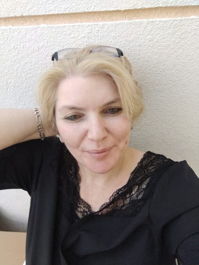 A medium shot captures a woman with blonde hair, wearing a black top with lace detailing around the neckline. She has on a silver bracelet on her left wrist and her left arm is bent, with her hand resting behind her neck. She is looking towards the camera with her head slightly tilted. She is wearing glasses pushed up on her head. The background is a textured, light-colored wall with a horizontal stripe. The scene appears to be outdoors, as suggested by the lighting and the textured wall, which is common in exterior architectural elements in regions like Al Mas'oudi, United Arab Emirates.