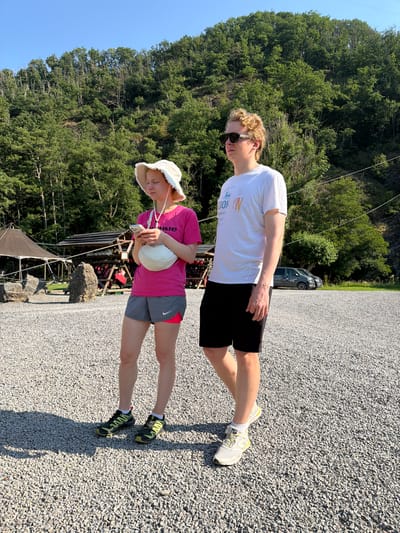 Two people are standing in a gravel parking lot. A woman with blond hair is wearing a pink t-shirt, gray shorts with neon pink trim, a white bucket hat, and black and yellow running shoes. She is holding a white fanny pack around her waist and looking at her phone. A man with blond, curly hair is wearing a white t-shirt, black shorts, sunglasses, and white and yellow running shoes. He is looking to the left. In the background, there is a dense green forest on a hillside. There are also some tents and structures set up, possibly for an outdoor activity or event. Two cars are parked in the distance. The sky is clear and blue. The location is identified as the city of Warisy, Belgium.