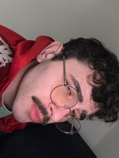 A man with dark curly hair and a mustache looks to the left while wearing round eyeglasses with yellow-tinted lenses. He sports thick eyebrows and a fair complexion. Part of a red hoodie, possibly Kappa brand, is visible around his neck. A dark fabric, perhaps a curtain, is in the background to the right. The background on the left is a plain light gray wall.