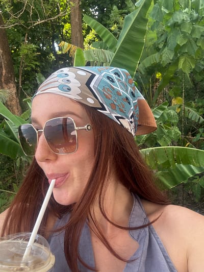 A red-haired woman wearing a patterned headscarf and sunglasses is seen sipping from a plastic cup with a straw. She is wearing a gray top. The background shows green tropical vegetation, specifically banana leaves. The location is Kamala, Thailand.