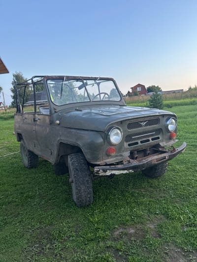 An olive-drab, open-top, vintage UAZ-469 jeep is parked on a grassy area in front of a rural house. The vehicle appears weathered and possibly abandoned, with rust visible on the roll cage and a dented front bumper. The jeep has its doors removed. In the background, a red house with white trim stands on a gentle slope, surrounded by trees and vegetation. The sky is clear and pale blue, indicating either early morning or late evening. The overall scene suggests a quiet, rural setting, likely in the village of Vereiki, Russia, given the architectural style and the prevalence of UAZ vehicles in the region.