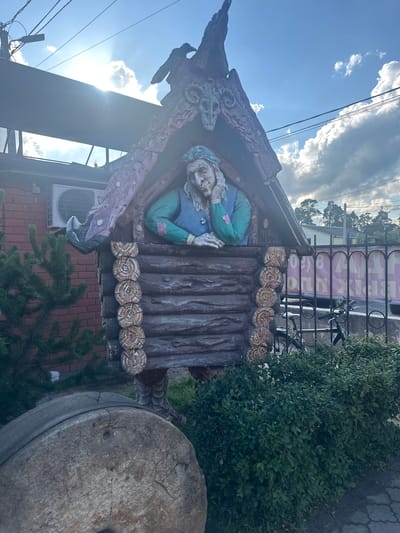The image depicts an outdoor scene in the city of Kablukovo, Russia, featuring a decorative statue of an old woman with a long beard, dressed in a blue vest and a green shirt, peeking out of a miniature log cabin. The statue has a skull-like ornament above it, and a dark bird silhouette on the roof. To the left of the statue is a brick building with an air conditioning unit and a pine tree. In front of the statue and to the right, there are green bushes and a black metal fence behind which bicycles are parked. A large, rough-hewn stone, possibly a millstone, sits in the foreground on the left. The sky is partly cloudy with the sun shining through.