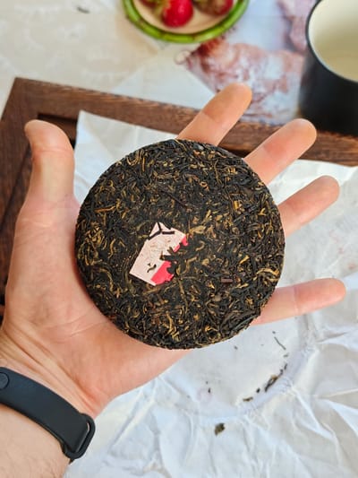 In a top-down shot, a person's left hand cradles a round, compressed cake of dark tea with golden flecks. A small, pink and white paper label, partially obscured by the tea leaves and featuring a red mark, is embedded on the surface of the tea cake. The tea cake is positioned above a crumpled white paper surface. In the background, out of focus, is a wooden tray, a white tablecloth with a subtle pattern, a black mug, and the rim of a green bowl containing red berries, likely strawberries. The scene suggests a domestic setting in Parėčča, Belarus, potentially a kitchen or dining area, where tea is being prepared or presented.
