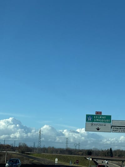 The image depicts a multi-lane highway scene under a clear blue sky with scattered white cumulus clouds along the horizon. In the foreground, traffic moves on the highway, with at least two vehicles visible: a dark gray sedan in the lower left moving left-to-right, and a red vehicle in the lower right, also moving left-to-right. A prominent green and white overhead road sign on the right directs traffic via route N 353 towards COLMAR and STRASBOURG, with a separate white panel indicating an exit for ENTZHEIM. A partial sign to the right also mentions STRASBOURG and PORT DE STRASBOURG. These locations are in France, specifically near Strasbourg in the Alsace region. The midground features a landscape with bare deciduous trees and numerous tall metal lattice power line towers stretching across the horizon. The perspective suggests the photograph was taken from a vehicle in motion.