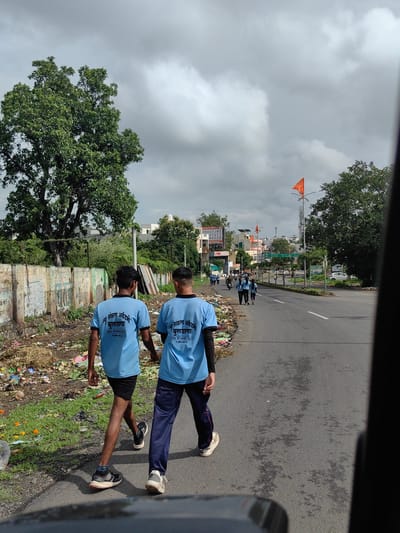 Two young men wearing blue t-shirts with "न्यू संकल्प अकॅडमी बुलडाणा" (New Sankalp Academy Buldhana) printed on them, walk side-by-side on a road in Buldhana, India. The t-shirts also have "Mo. 8766451267" and "मा. पश्चिम वर्ध" printed on them. One man wears black shorts and the other wears dark blue track pants. They are walking away from the camera, on the right side of the road. The road has a white lane marking. To the left of the road, there is a wall covered in graffiti and piles of garbage. Beyond the wall, there is lush green vegetation and a large tree. In the background, there are buildings, streetlights, and an orange flag on a pole. The sky is overcast with dramatic clouds. The overall scene suggests an urban environment with some signs of disarray and activity.