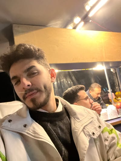 A man with dark hair and a beard is looking at the camera. He is wearing a vest and a light-colored jacket. Behind him is a reflective surface showing another man with glasses eating something. A column is next to the reflective surface and there is a row of lights to the side.