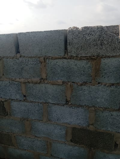 A medium shot captures a wall constructed from grey cinder blocks, laid in horizontal rows with visible tan mortar. The top edge of the wall is uneven, with some blocks broken or protruding. Above the wall, a pale sky with scattered white clouds is visible, suggesting an outdoor setting. The scene appears to be in Dutsen Alhaji, Nigeria.