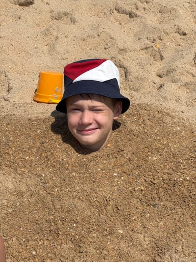 A young boy is buried in the sand up to his neck on a beach in Vityazevo, Russia. He is wearing a bucket hat with red, white, and blue sections. To his left, an orange plastic bucket is partially visible. The sand is light brown and textured, with some pebbles and shells mixed in. The boy is smiling and has his eyes partially closed, suggesting he is enjoying the experience. The scene appears to be a sunny day, as indicated by the lighting and shadows.