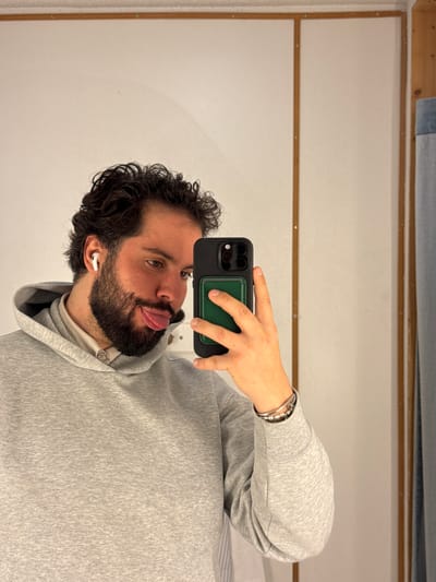 The image displays a male individual, appearing to be an adult, taking a mirror selfie indoors. He possesses dark, curly hair, a full beard, and a mustache. A white wireless earbud is visible in his left ear. He is wearing a light grey hooded sweatshirt over a light-colored collared shirt. On his right wrist, he wears several bracelets, including silver-toned bands and a string of white beads. With his left hand, he holds a smartphone featuring a dark case and a green accessory attached to its back, partially obscuring the lower right side of his face. The individual is looking towards his right and has his tongue slightly extended. The background consists of a plain, light-colored wall. Visible elements include a horizontal wooden strip at the top and a vertical wooden frame on the right side of the wall. A fragment of light blue fabric is discernible on the far right edge of the frame. The specific location within the city of Gondomar, Portugal, is not identifiable from the provided image.