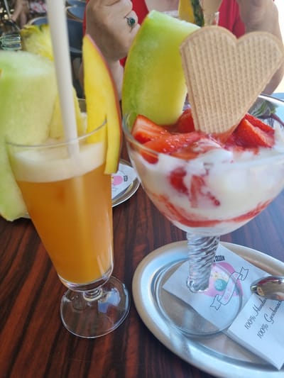 The image depicts a table at a cafe, possibly in Saarbrücken, Germany. There is a dessert glass with strawberries, ice cream and a heart-shaped wafer cookie. The glass is partially obscured by a slice of melon. Beside it, there's a glass of orange juice with melon slices and mango garnish. A silver tray is visible, holding napkins, a logo of "Heiss Heiss Eis", and a spoon. The table is wooden and has a dark wood grain. A woman's hand with a ring is visible near the dessert glass. The scene suggests a relaxing moment at a cafe.