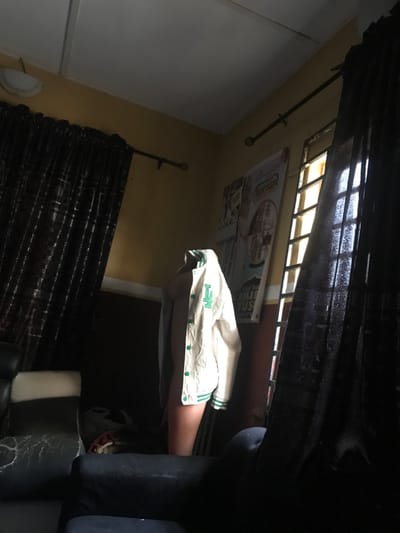 The image depicts an indoor scene within a room located in Oba-Ile, Nigeria. A standing human figure is positioned centrally, partially obscured by shadows, wearing an open, cream-colored varsity-style jacket with green trim, green buttons, and a prominent green "LA" emblem on the left chest. The figure's head is not visible, appearing to be covered or obscured by the jacket's hood, and bare legs of light skin tone are visible below the jacket.
The room features light brown/tan walls, a white ceiling with a visible beam, and a white decorative light fixture in the upper left corner. Two windows are present; the left is mostly covered by a dark, checkered curtain, while the right window, partially covered by a matching curtain, reveals bright exterior light filtered through vertical metal grilles. Dark curtain rods with finials are mounted above both windows. Two light-colored posters are affixed to the wall between the windows. Furniture includes a black armchair or sofa in the left foreground with visible wear and a white armrest, and a dark-colored sofa armrest in the right foreground. The room is dimly lit, with strong backlighting from the right window creating deep shadows. No discernible actions or interactions beyond the figure standing are visible.