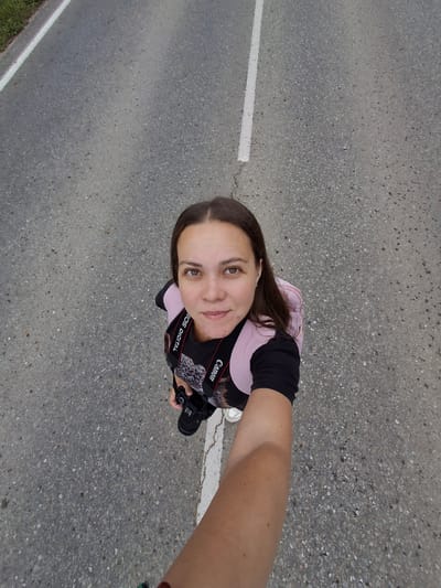 A medium-skinned woman with long brown hair is captured in a selfie taken from a high angle on an asphalt road. She is wearing a black t-shirt with a graphic print and a pink backpack. A camera with a Canon strap is visible, held in her right hand. She is standing on the asphalt, which is divided by a white line. To the left of the road, there is a strip of green grass. The overall setting suggests an outdoor location, possibly in the city of Garevaya, Russia, given the presence of the road and the natural elements.