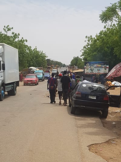 The image shows a street scene in Dabawa, Nigeria. The road is lined with green trees and several cars, including a blue sedan, a red hatchback, and a white truck. Several people are walking on the road. A billboard displaying photos of political figures is visible in the background.