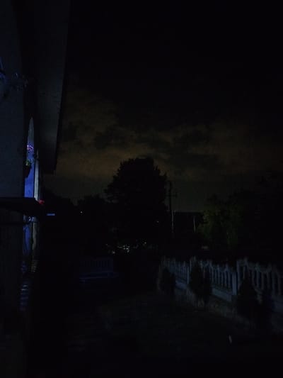 This image depicts a dark, outdoor scene at night, likely in Nimoreni, Moldova. The foreground is dominated by a dark, textured structure on the left, possibly a building facade or a porch roof, casting a significant shadow. A sliver of light, possibly from an artificial source, illuminates part of this structure and a glimpse of what appears to be greenery or plants.
Beyond this structure, the scene opens to a yard or garden area. A dark, silhouetted tree with dense foliage stands prominently in the mid-ground, against a faintly illuminated sky. The sky itself is obscured by clouds, which are catching some ambient light, creating a muted glow.
Further into the background, a decorative fence is visible, with an ornate, balustrade-like design. Several small, conical evergreen trees are planted in front of this fence, also silhouetted against the dark. Beyond the fence, the outlines of other trees and possibly structures are discernible, though heavily obscured by darkness. The ground appears to be covered in grass or dirt, with fallen leaves possibly present. There are no visible people or animals in the image. The overall impression is one of a quiet, tranquil night scene in a residential or garden setting.