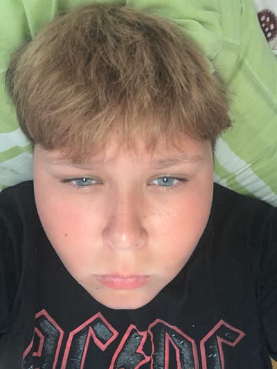 This is a close-up, head-on, overhead, indoor photograph of a young person with light brown, tousled hair and blue eyes, looking directly at the camera with a neutral expression. The person is wearing a black t-shirt with a red and grey graphic print. They are lying down on a green and white striped surface, likely a bed or couch, with part of a patterned brown and white fabric visible in the upper right corner of the frame. The lighting appears to be natural or soft artificial light, casting a subtle shadow on the person's face. The image is focused on the upper body and face of the individual, with the background slightly out of focus. The location is Vejprty, Czechia.