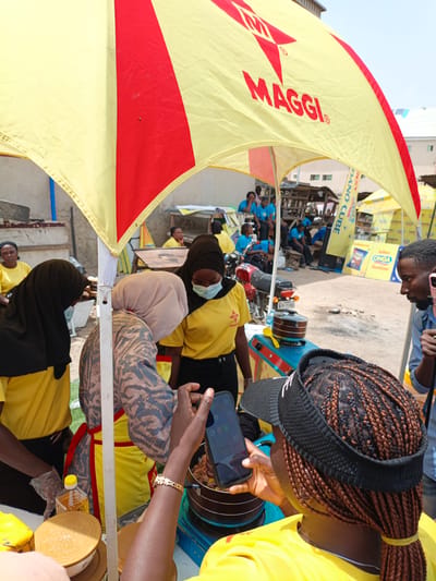 The image depicts a bustling outdoor scene, possibly a market or event, in Tarauni, Nigeria. The foreground is dominated by a large, brightly colored yellow and red umbrella, advertising "MAGGI". Underneath the umbrella, several people are engaged in an activity.

In the center, a woman wearing a yellow shirt and a headscarf, possibly a vendor, is attending to a cooking pot on a small stove. Another person, also in a yellow shirt and headscarf, is filming the cooking with a smartphone. To the left, another woman in a yellow shirt and black headscarf stands, looking towards the cooking area. Further in the background, a man in a blue shirt is also present.

The setting appears to be a street or open area with buildings and stalls visible in the distance. There are other tents or umbrellas in the background, suggesting a larger event or market. A motorcycle is parked to the left of the cooking station. The lighting suggests it is daytime, likely sunny. The overall atmosphere seems active and engaged.

Visible text includes "MAGGI" on the umbrella, and on banners in the background, "DANO CUBE", "Saborn", and "ONGA Dandano". A license plate on the motorcycle reads "SR-75 800".