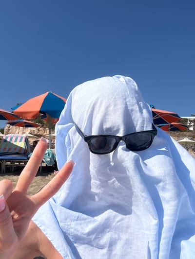 The image depicts a person on a sandy beach in Peyia, Cyprus, seemingly covered by a white sheet with black sunglasses placed over the covered face, creating a "ghost" appearance. The person is holding up two fingers in a peace sign. In the background, beach umbrellas in orange and blue are visible, along with beach chairs and a rocky hillside. The sky is clear blue.
