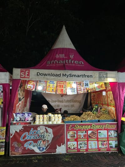 A street food stall at night features a Smartfren-branded tent and a sign promoting "Download MySmartfren". Colorful menus display various food items like "Teruk Ras" and "Es Dogan". A large sign reads "L-Sagara Street Food", listing burgers, grilled sausages, and ice cream rolls. Ice cream cones are stacked nearby. The stall sells "Ecer Buah Lokal Malang - Jeruk RGL Jeruk Lemon" (Local Malang Fruit). A person wearing a white cap is visible inside the stall.