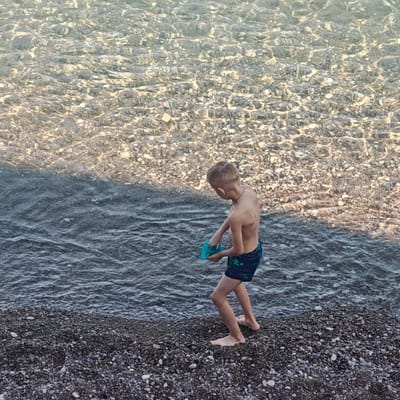 A young boy stands on a pebble beach at the water's edge, holding a blue toy. The water is clear and shallow, with sunlight reflecting off the pebbles on the seabed. The boy is wearing dark swim trunks with a green palm leaf pattern. The image captures a moment of leisure and play by the sea, suggesting a location in a coastal area.