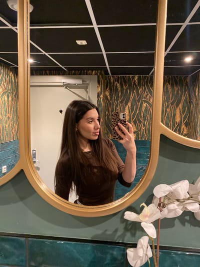 A woman is taking a selfie in a bathroom mirror. The mirror has a gold frame and is mounted on a wall with various patterned tiles. To the right of the mirror, there is a fake orchid. Behind the woman in the mirror is a white door. The location is Uppsala, Sweden.