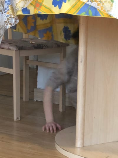 The scene is an interior view, located in Bouxwiller, France, depicting a low-angle perspective under and around a light-toned wooden table. A yellow tablecloth with a repeating pattern of blue floral elements and orange grid lines covers the table's surface. A section of a white, open-weave lace or crocheted tablecloth is partially visible on the left. To the left of the table stands a light-colored wooden chair with a padded seat upholstered in a mottled brown fabric; a small, irregular light-colored fragment is situated on the seat cushion. A child, attired in light grey clothing, is observed on the light brown wooden floor, positioned beneath the table. The child's left arm is extended forward and to the left, with the fingers of their hand splayed and resting flat on the floor surface. A portion of the child's head, showing dark hair, is discernible behind the table's right-side vertical support. The background reveals a white wall and a light-colored wooden baseboard along the floor.