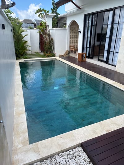 The image depicts an outdoor swimming pool situated in Ungasan, Indonesia, bordered by a white stone edge. The pool's water has a blue-green hue. One side of the pool is flanked by a wooden deck and a row of white stones. Adjacent to the deck is a building with black-framed windows and doors. An outdoor seating area with a woven design can be seen through an arched opening. A tropical landscape with palm trees and other greenery is visible. The sky is blue with scattered clouds. The image is taken from an elevated perspective.