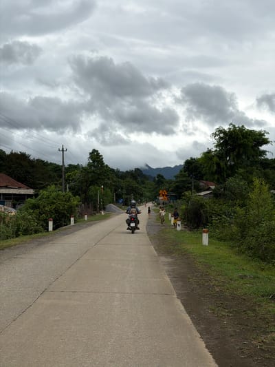 The image depicts a rural road scene in Kim Ngân Commune, Vietnam, under an overcast sky with dark, dramatic clouds. A paved road runs through the center of the frame, bordered by green vegetation and trees on both sides. In the foreground, a person is riding a motorcycle away from the viewer. Several children are visible walking on the side of the road further ahead. On the left side of the road, a house with a tiled roof is partially visible, surrounded by dense foliage and a utility pole with wires. On the right side of the road, a pile of gravel and more trees are present. The overall atmosphere is subdued, likely due to the cloudy weather.