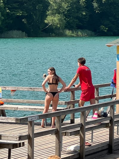 In Vilnius, Lithuania, on a sunny day, a young woman in a black bikini stands on a wooden pier, facing away from the camera with her hands on her hips. Beside her, a young man in a red t-shirt and red shorts leans on the pier railing, looking out at the water. The turquoise water of a lake stretches out before them, bordered by a dense green forest. Two orange buoys are visible in the water. The pier is constructed of weathered wooden planks and railings, with a few items resting on it, including a blue and red life jacket, a pink and white striped object, and a pair of shoes.