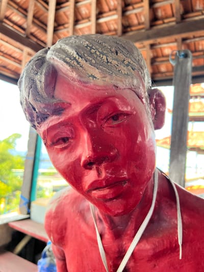 The image shows a close-up, low-angle shot of a red statue of a young person's head and shoulders. The statue has rough, textured gray hair and a red face with carved features. A white cord is visible around the neck of the statue. In the background, the wooden structure of a building with an orange tiled roof is visible, along with blurred greenery and what appears to be a distant structure. The lighting suggests an outdoor or semi-outdoor setting during the day. This statue is located in the village of Tejakula, Buleleng Regency, Bali, Indonesia.