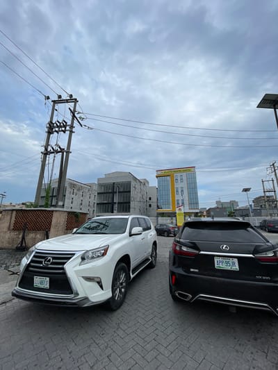 This media file depicts a street scene in Moba, Nigeria, with a focus on two parked vehicles and a building. The primary subjects are a white Lexus SUV in the foreground, angled towards the viewer, and a black Lexus SUV behind it. The white SUV has its license plate, "JJJ-487 JF", clearly visible, which indicates "LAGOS". The black SUV's license plate, "APP-95JR", also indicates "LAGOS" and the model "RX350".
The scene includes architectural elements, notably a modern building with a yellow and glass facade, identified by signage as "ATLANTIS PEDIATRIC HOSPITAL". Several utility poles with numerous wires are prominent in the background and foreground, suggesting an urban or semi-urban environment. The ground is paved with brickwork.
The sky is overcast with heavy clouds, indicating a cloudy or potentially rainy day. The time of day is indeterminate due to the lighting, but the overall atmosphere is neutral. There are no people visible in the frame. The composition is such that the image is oriented vertically, as if taken with a phone held upright. The prominent Lexus vehicles and the hospital building are the main points of interest.