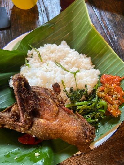 A culinary scene captured in Batu, Indonesia, presents a traditional meal served on a white plate with a blue rim, lined entirely with vibrant green banana leaves. The primary subject is a whole, deep-fried duck, exhibiting dark brown, crispy skin and visible bone structure, positioned prominently on the lower left of the plate. To the right of the duck, a generous portion of white, fluffy rice is mounded, with several thin, green stems, likely from a leafy vegetable, embedded within and atop it. Adjacent to the rice, a serving of bright green, glistening stir-fried leafy vegetables, resembling water spinach, is arranged. Further right, two distinct types of sambal are present: one is a smooth, reddish chili paste, while the other is a coarser preparation featuring finely chopped red and green chilies, shallots, and possibly other components. A single red chili pod is visible tucked beneath the fried duck, and scattered small white specks (possibly rice grains) rest on the banana leaf. The entire meal is set on a dark brown wooden table, which displays prominent grain and texture. In the blurred background at the upper left, a yellow cylindrical object and a dark, rectangular item are partially visible.