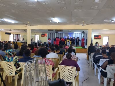 The interior of a church located at coordinates -6.828, 37.658 in Morogoro, Tanzania. The church is filled with people seated in rows of yellow plastic chairs facing a stage. There are approximately 75 people in attendance. In the front of the room, approximately 15 people are standing on a stage, singing, and performing on instruments. They are wearing dark suits, dresses, and gowns. The ceiling is white with square tiles, and there are ceiling fans. The floor is beige tiled. The walls are painted a light yellow. There are a number of windows on the walls. There are two large flat-screen TVs displaying images. The air is warm and still. There is a sense of peace and quiet.