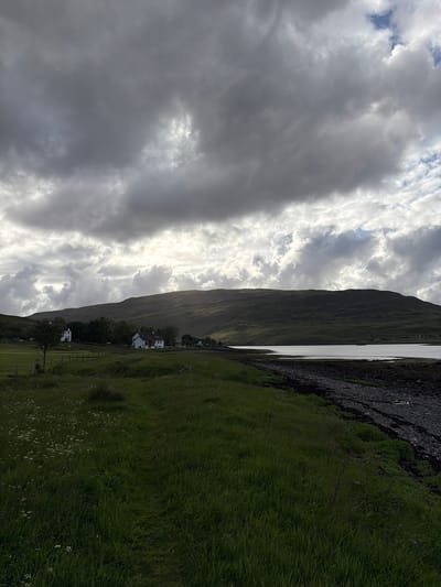 A wide shot captures a coastal landscape under a dramatic, cloud-filled sky. The foreground is dominated by a grassy field with scattered white wildflowers and a faint path leading towards the coast. To the left, a few houses are visible nestled against a tree line, with a fence separating the field from the houses. Beyond the houses, rolling green hills form the backdrop, sloping down to meet a calm body of water. The water's edge is a rocky shoreline. The sky is a mixture of dark, heavy clouds and areas where sunlight breaks through, casting a diffused light on the landscape. The scene appears to be located in Sconser, United Kingdom.