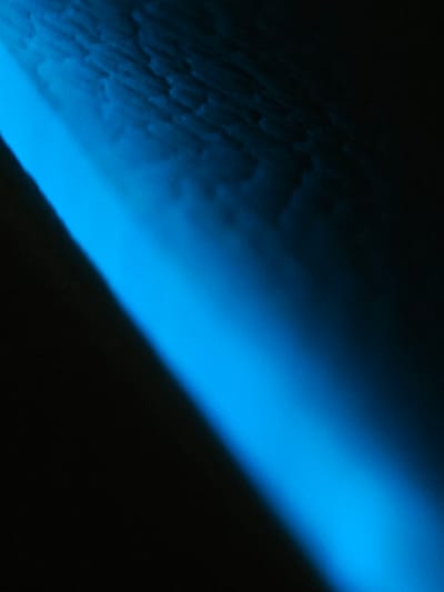 The image depicts a dark environment illuminated by a strong, diagonal beam of bright blue light. The light casts a gradient, starting intensely bright and fading into darkness. The upper portion of the image shows a textured surface, possibly a wall or ceiling, with a pattern of raised, rectangular elements, appearing somewhat rough and uneven. This textured surface is partially illuminated by the blue light, revealing subtle shadows and highlights. The rest of the scene is shrouded in deep shadow. There are no visible people, animals, or distinct objects other than the implied surface and the light source. The image does not provide any specific visual cues to identify a location in the city of Bo Y, Vietnam.