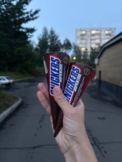 In this media file, two Snickers ice cream cones are held by a person's hand against an outdoor backdrop. The cones are chocolate-covered and feature the prominent "SNICKERS" logo in blue and white, with Russian text below. The text on the packaging mentions "МОРОЖЕНОЕ" (ice cream), "АРАХИСОВОЕ" (peanut), and "КАРАМЕЛЬЮ" (caramel), indicating the flavor profile and composition. The setting appears to be urban, with a blurry apartment building visible in the background, suggesting a residential area. Trees are also present, contributing to an outdoor, possibly park-like or street-side atmosphere. The lighting suggests it is either early morning or late evening, given the soft, diffused light and the blueish hue of the sky. The ground appears to be asphalt, possibly wet from recent rain, indicated by darker patches and reflections. The image is shot from a slightly high angle, looking down towards the ice cream cones. The overall scene conveys a casual moment of enjoying a treat outdoors.