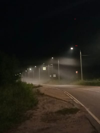 The image depicts a nighttime scene on a rural road in Voskresenskoye, Russia, characterized by heavy fog. A paved road curves to the right, with a double white line marking the center. To the left of the road is a dirt shoulder with sparse grass and vegetation. Further left, a dense line of dark green foliage lines the edge of the road.
Several streetlights line the road, their illumination diffused by the fog, creating hazy glows. Two of these streetlights are equipped with flashing red lights at their tops, and another has a square yellow sign with a pedestrian crossing symbol illuminated. A traffic sign with a red "K" symbol on a white background is also visible on a pole to the right of the road. The sky is dark, with a faint red light visible in the upper portion of the frame, possibly an aircraft warning light. The overall atmosphere is somber and slightly eerie due to the fog and darkness. There are no people or vehicles visible in the image.