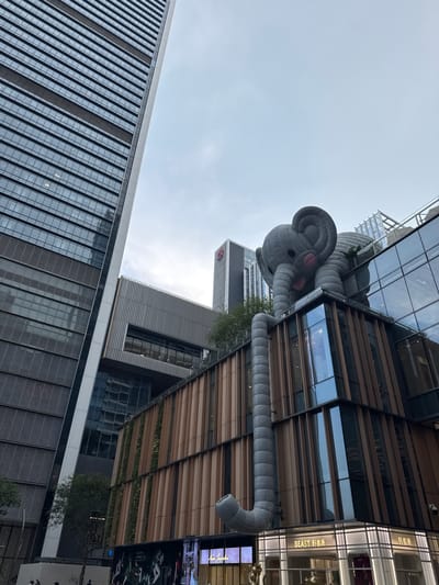 An upward-angled, eye-level shot captures a cityscape in the Nanshan District, China. The focal point is a large, soft, gray, inflatable elephant sculpture perched atop a modern building. The sculpture's trunk extends downwards, ending in a curve. The building it sits on has a contemporary design, featuring alternating bands of glass and wooden slats. Adjacent to this building, a tall, modern skyscraper with a glass facade stretches upwards. Patches of greenery are visible, suggesting vertical gardens or landscaping on the buildings. The sky is overcast, casting a soft, diffused light over the scene. The angle of the shot emphasizes the height and scale of the buildings and the inflatable artwork.