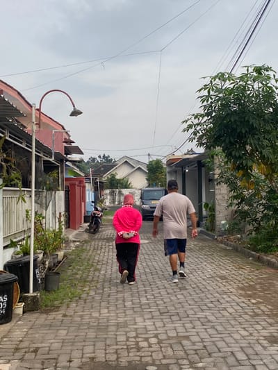 The image shows a man and a woman walking down a narrow cobblestone street in Piyungan, Indonesia (latitude: -7.827, longitude: 110.483). The man, wearing a light grey t-shirt and blue shorts, walks in front, while the woman, wearing a pink hijab and black pants, follows behind. The street is lined with houses on both sides, and there is a small motorcycle parked on the left side. There is a tree and some plants on the right side. The sky is overcast, and the environment is quiet. There are power lines running over the street. The man and woman are not interacting. The man is looking ahead, and the woman is looking down at her feet. The street appears to be residential, and there are no other people in the vicinity.