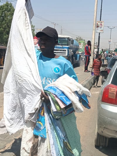 This image captures a street scene in Hotoro South, Nigeria, under bright daylight. In the immediate foreground, a young male, appearing to be a vendor, with dark skin and a neutral expression, stands facing the camera. He wears a black baseball cap and a light blue short-sleeved t-shirt with a white "N" logo on the left chest and "MSC" text below it. He holds a large, crumpled bundle of white fabric in his left arm, extending upwards. In his right hand, he grasps a stack of folded, multicolored textiles, likely sheets or fabrics, which extend downwards to his side. A ring is visible on his right ring finger.
To his immediate right, the rear portion of a silver sedan is visible, parked or moving slowly. Behind the primary subject, a white DAF truck, possibly a cement mixer with a blue drum and "ALLAH" written on its front, is present in the midground. Further back and to the left, part of a yellow auto-rickshaw (keke Napep) is discernible. A political poster, partially obscured, depicting a man in a red cap with "GOVERNOR" text, is visible behind the vendor's left shoulder.
In the midground to the right, a male in a red and black tracksuit stands by a utility pole, appearing to be on a mobile phone. A young child in a red and black striped t-shirt and dark pants stands nearby, holding a purple object. The street beyond is populated with other pedestrians and vehicles, including other cars and motorcycles. A second political poster featuring a man in a white cap is affixed to the utility pole. The sky is clear and bright, indicating a sunny day. The scene depicts a bustling roadside environment.