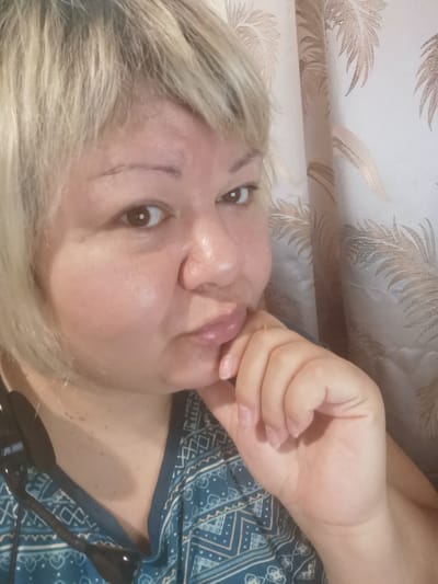 A fair-skinned woman with blonde hair is looking into the camera with her right hand resting on her chin. She is wearing a teal and white patterned shirt. The background consists of a beige curtain with a palm leaf pattern. The image appears to be a selfie taken indoors. There is no visible indication of the location being the city of Voronezh, Russia.