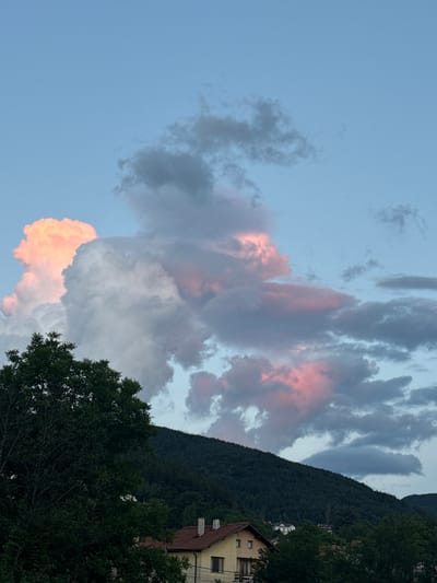 The image depicts a landscape scene with a focus on the sky and a mountainside. The sky is filled with dramatic clouds, some of which are tinged with pink, suggesting it is either sunrise or sunset. Dark green foliage, likely trees and a mountainside, occupies the right side of the frame. A building, possibly a house, is visible nestled on the mountainside, with its roof and upper floors showing. The overall mood is serene and natural, with the colors of the sky creating a visually appealing contrast with the darker elements of the landscape. The image appears to be taken in Rudartsi, Bulgaria. There is no visible text in the image.