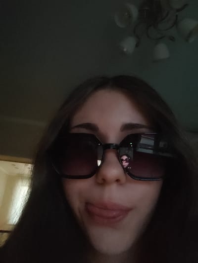 A selfie taken from a low angle shows a young woman wearing large, dark sunglasses. Her mouth is open, and her tongue is sticking out slightly. Her dark hair frames her face. In the reflection of her sunglasses, another person can be seen wearing similar sunglasses, with a blurred background that appears to be an indoor setting with a chandelier. The context of Svietlahorsk, Belarus is not evident from the image itself.