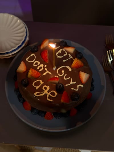 A birthday cake with the inscription "Don't age" is displayed on a dark blue plate. The cake is covered in chocolate frosting and decorated with strawberries, blueberries, and white icing. A lit birthday candle is placed in the center of the cake. To the left of the cake, a stack of white plates with a blue rim is visible. To the right of the cake, two gold forks are partially visible. The scene appears to be indoors, with a dimly lit background featuring purple and dark hues. The image does not provide any specific details that would allow for the determination of its location in Chang Phueak, Thailand.